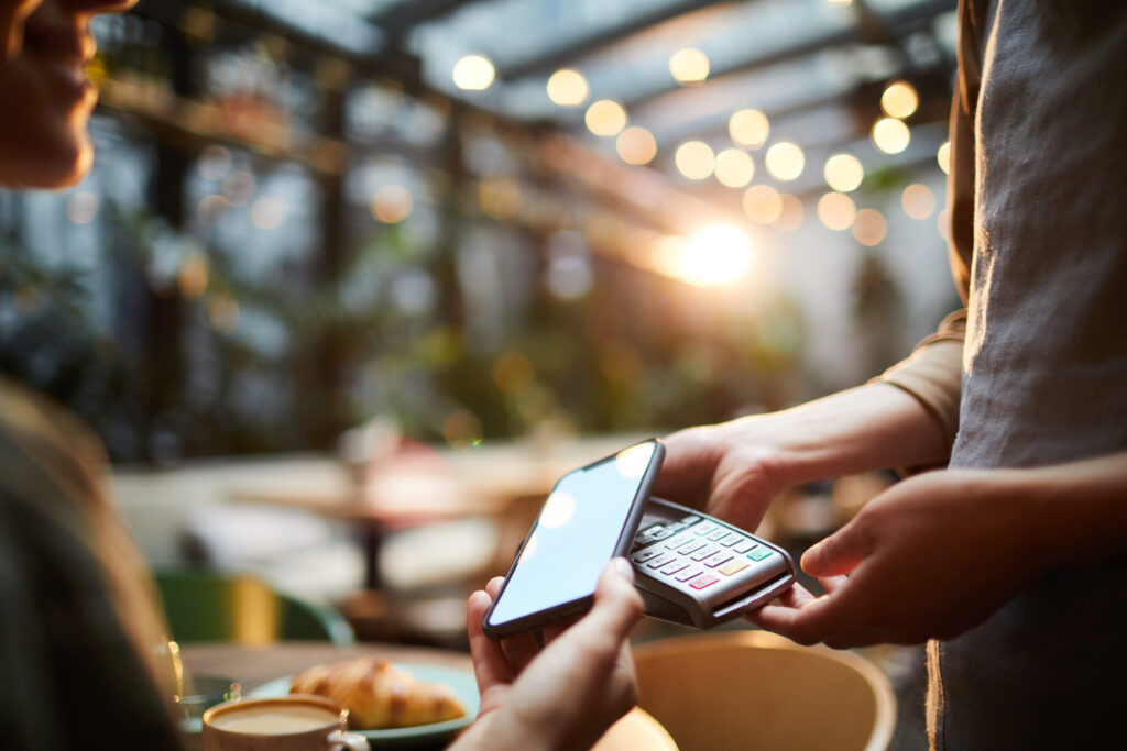 Close-up of unrecognizable customer using smartphone for online payment and putting device to terminal in modern cafe